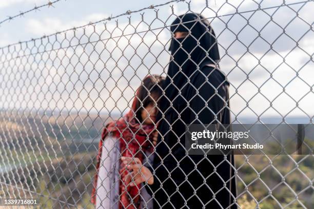 refugee woman and daughter standing behind a fence - burka stock pictures, royalty-free photos & images