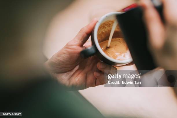 over the shoulder asian chinese male barista pouring froth milk on coffee cup prepared coffee latte art at bar counter - koffiemachine stockfoto's en -beelden
