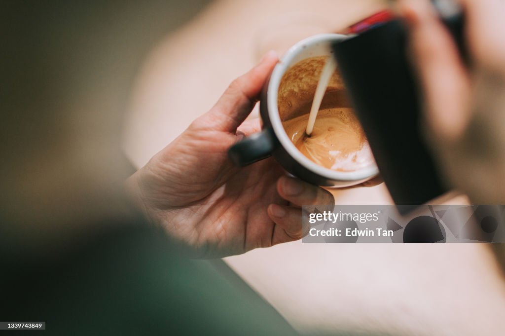 Over the shoulder asian chinese male barista pouring froth milk on coffee cup prepared coffee latte art at bar counter