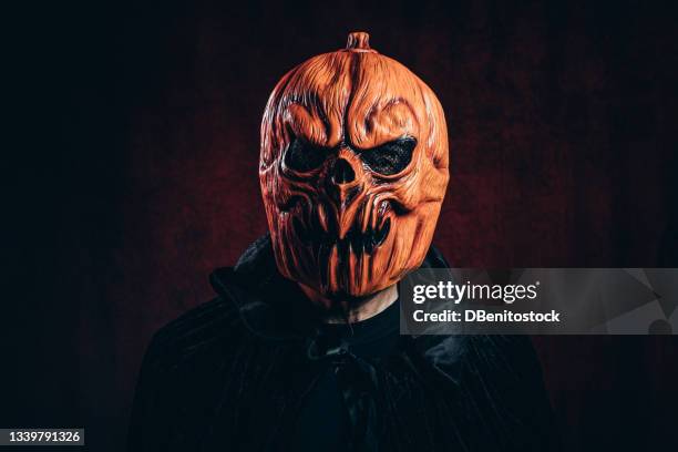 portrait of person in scary halloween pumpkin mask with black cape, in dark room with victorian velvet background and red light. - ugly pumpkins stock pictures, royalty-free photos & images