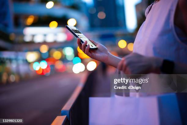 young woman carrying a paper shopping bag, managing online banking on smartphone in city street against illuminated street lights. tracking and planning spending. transferring money, paying bills, checking account balances. smart banking with technology - e commerce stock pictures, royalty-free photos & images