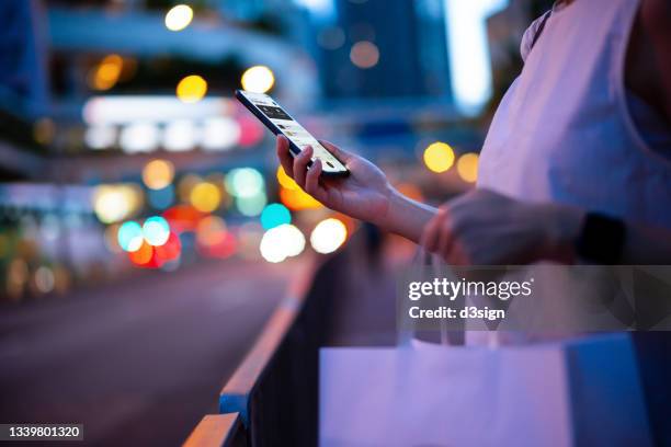 young woman carrying a paper shopping bag, managing online banking on smartphone in city street against illuminated street lights. tracking and planning spending. transferring money, paying bills, checking account balances. smart banking with technology - chinese young adults shopping imagens e fotografias de stock