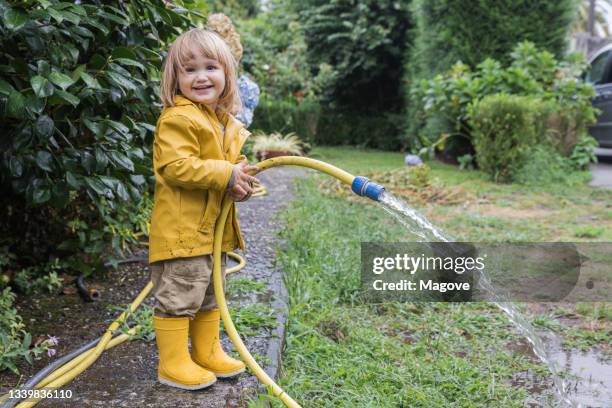 side view of cute little child wearing yellow raincoat and rubber boots watering garden from hose after rain and looking at camera - hose stockfoto's en -beelden