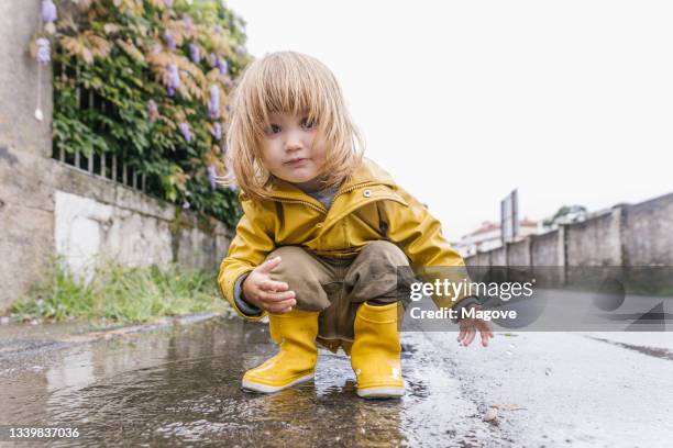 low angle of adorable little child in yellow raincoat playing in puddle and splashing water while entertaining after rain on street looking at camera - water repellent stock pictures, royalty-free photos & images