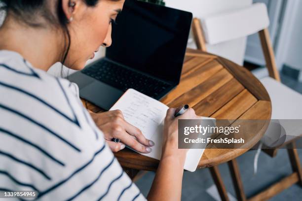 anonymous woman sitting at a wooden table, using her laptop and taking notes - writing a list stock pictures, royalty-free photos & images