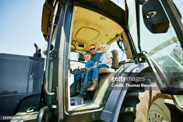 Medium wide shot of farmer sitting with grandchildren in cab of tractor during wheat harvest