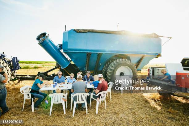 Wide shot of multigenerational farm family gathered for meal in wheat field during summer harvest