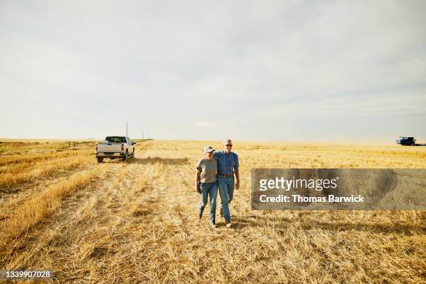 Wide shot of smiling embracing farm couple walking through cut wheat field during summer harvest