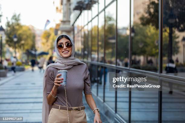 mid adult woman with cup of coffee on city street in morning. - arab woman walking stockfoto's en -beelden