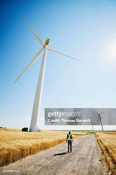 wide shot of engineer walking down service road to inspect wind turbines in wheat field on summer afternoon - mill worker stock pictures, royalty-free photos & images