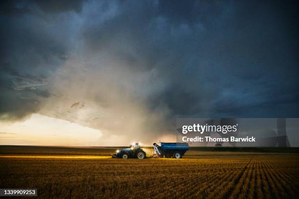 wide shot of tractor pulling grain cart through cut wheat field ahead of storm clouds during summer harvest - farm ストックフォトと画像
