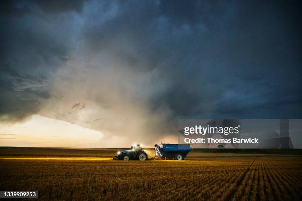 wide shot of tractor pulling grain cart through cut wheat field ahead of storm clouds during summer harvest - farm stockfoto's en -beelden