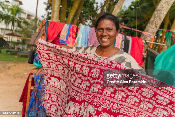 local sri lankan indian woman senior positive smiling cheerful woman, the small business owner, demonstrating textile in her open-air clothing and souvenirs store on the beach. - sri lankan culture stock pictures, royalty-free photos & images