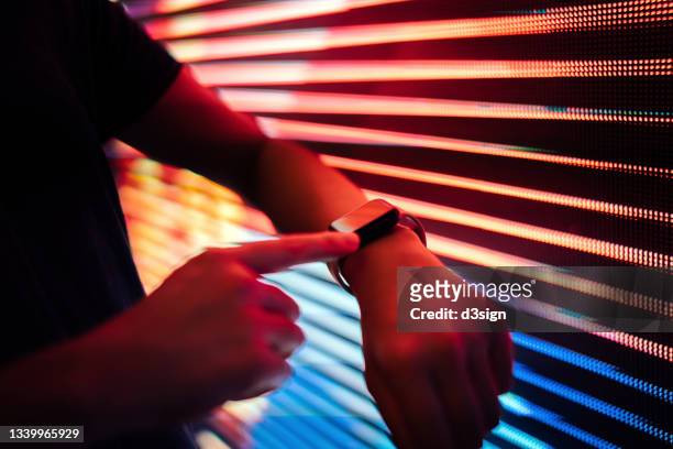 close up of young woman using smart watch in front of illuminated and multi-coloured led display screen in the city - montre connectée photos et images de collection