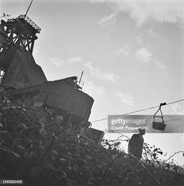 Mine worker looks up at an aerial ropeway transporting tin ore from the pit head to the dressing plant at East Pool tin mine near Camborne in...