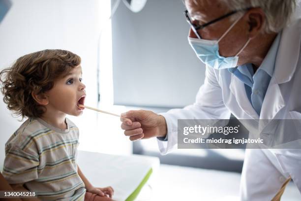 pediatrician with protective face mask examining boy's throat in his office at medical clinic - throat exam stock pictures, royalty-free photos & images