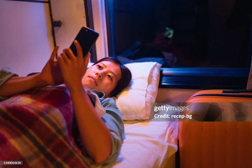 Junge weibliche Reisende, die bequem in einem Bett im Zugabteil liegt und nachts ein Smartphone benutzt