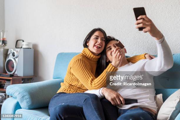 latin-paare sind auf dem sofa in ihrem wohnzimmer, machen ein selfie und essen schokolade - couple chocolate stock-fotos und bilder