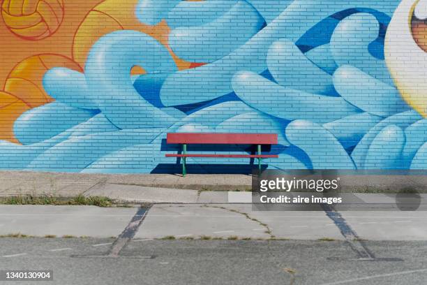 an empty red bench on a sidewalk with a graffitied wall as background. an empty red bench in front of a blue graffitied mural on a sidewalk. - street fotografías e imágenes de stock