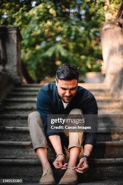 happy man tying shoelace sitting on steps in city in summer - lacet de chaussures photos et images de collection