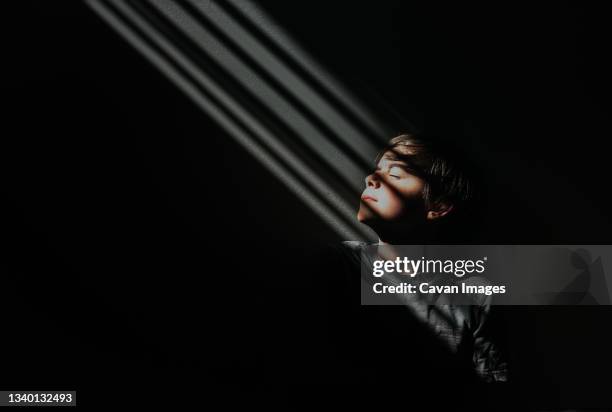 young boy sitting in a patch of patterned light in a dark room. - child eyes closed stock pictures, royalty-free photos & images