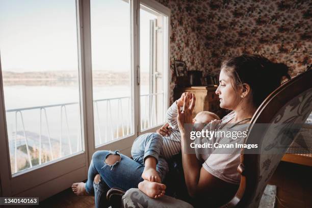 mother breastfeeding baby on chair in vintage room looking out on lake - latch stock pictures, royalty-free photos & images