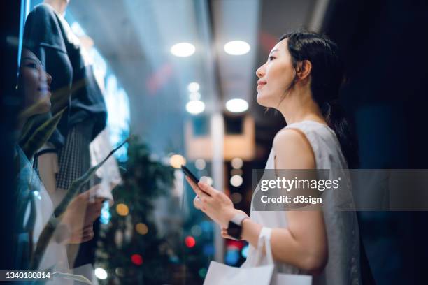 young asian woman carrying a paper shopping bag, checking her smartphone while standing outside a boutique looking at shop window in the evening in city - veleiding stockfoto's en -beelden