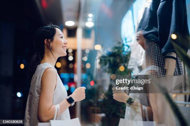 smiling young asian woman carrying a paper shopping bag, standing outside a boutique looking at shop window in the evening in city - store window stock pictures, royalty-free photos & images