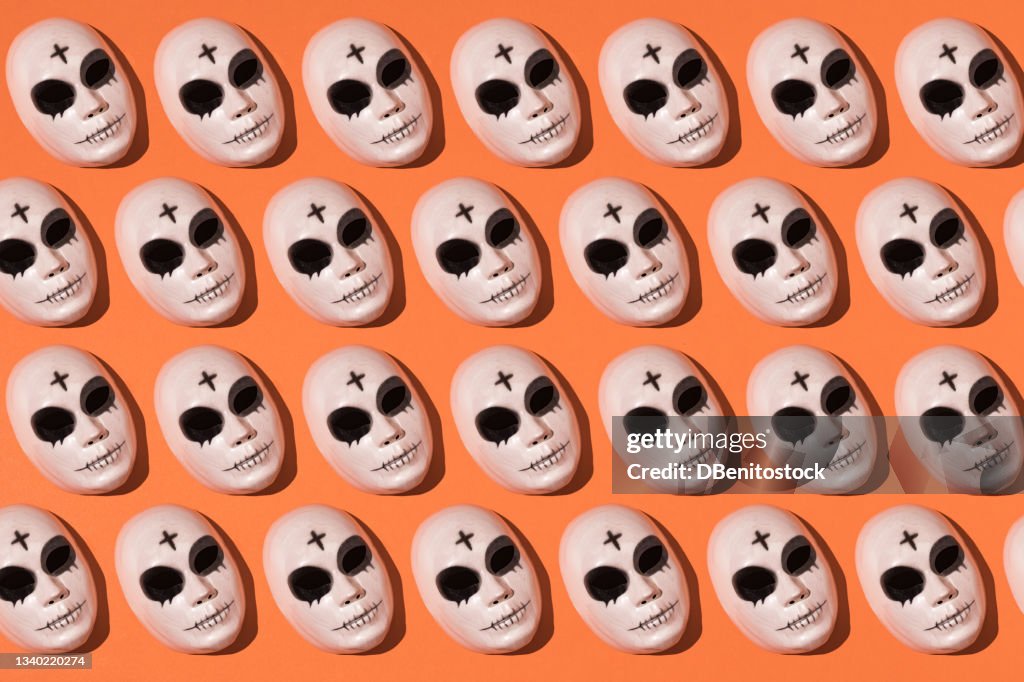 Repetitive halloween killer mask pattern with a cross on the forehead and pronounced dark circles on orange background. Halloween celebration concept.