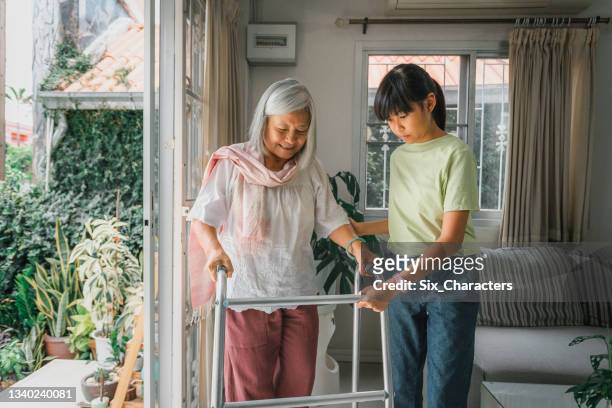 asiatische enkelin hilft ihrer großmutter patienten, mit mobilitätsläufer im wohnzimmer zu hause zu gehen - assisted living stock-fotos und bilder