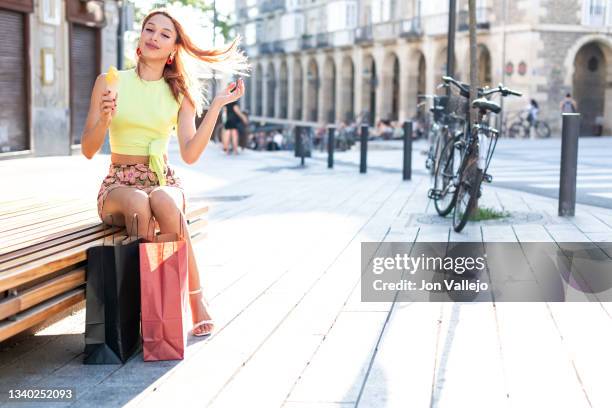 preciosa mujer sentada en un banco de madera tocandose el pelo mientras se esta comiendo un helado al lado de sus bolsas de la compra. - mujer sentada stock pictures, royalty-free photos & images