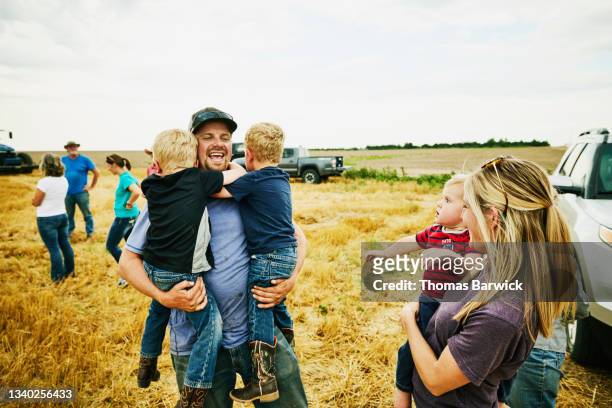 Medium wide shot of laughing farmer holding young sons in cut wheat field during summer harvest
