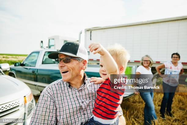 medium shot of laughing farmer holding grandson during family meal in wheat field during harvest - american family stockfoto's en -beelden