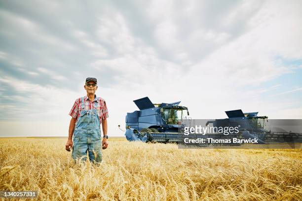 Wide shot of smiling farmer standing in wheat field in front of combines during summer harvest