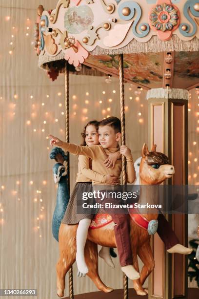 funny kids teen friends a little girl and a little boy ride together on a merry-go-round with horses on the background of garlands and a christmas tree in an amusement park in the new year - carousel horse stock pictures, royalty-free photos & images