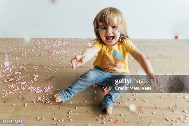 from above of cheerful infant child sitting on floor and throwing up colorful sprinkles and confetti while having fun celebrating in nursery - messy playroom stock-fotos und bilder