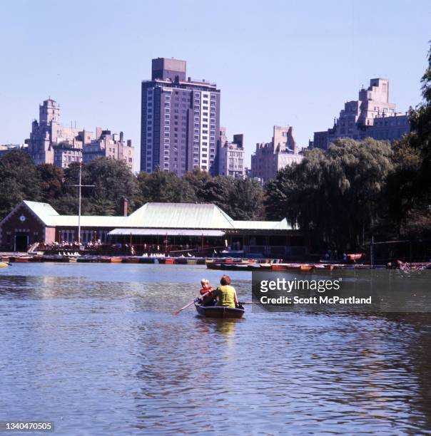Two people paddle in a rowboat on Central Park lake, New York, New York, September 1969. Visible is the Loeb Boathouse on the lake's far shore.
