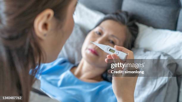 young asian female doctor holding thermometer taking temperature for mature woman at home - taking temperature stock pictures, royalty-free photos & images