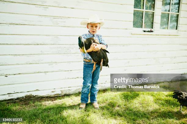 Wide shot portrait of young boy holding duck outside of chicken coop on farm on summer morning