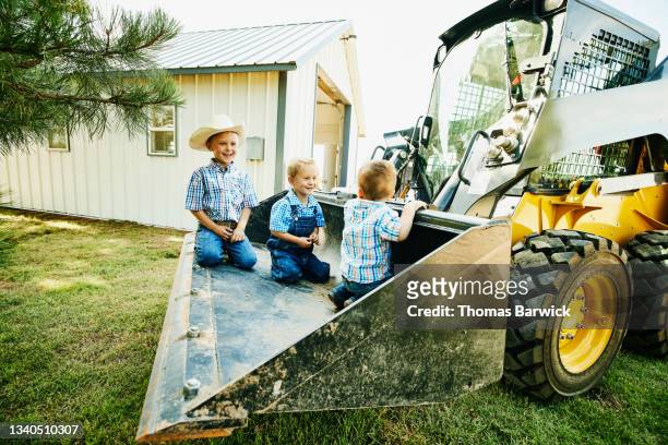 Wide shot of smiling young brothers riding in bucket of skidsteer on farm