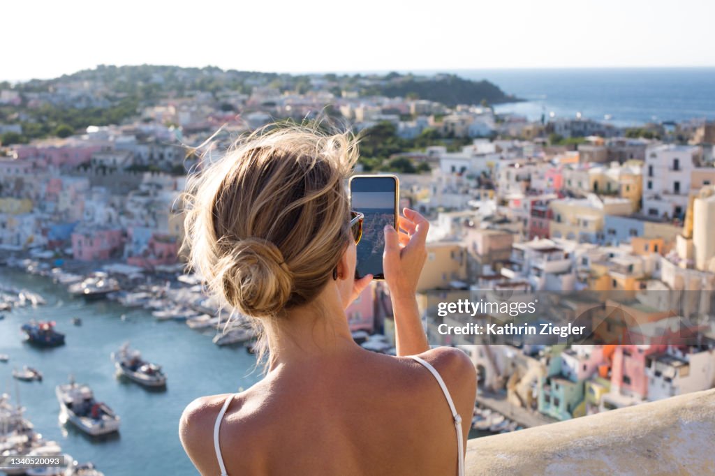 Woman taking pictures of the idyllic port of Corricella, Procida Island, Italy