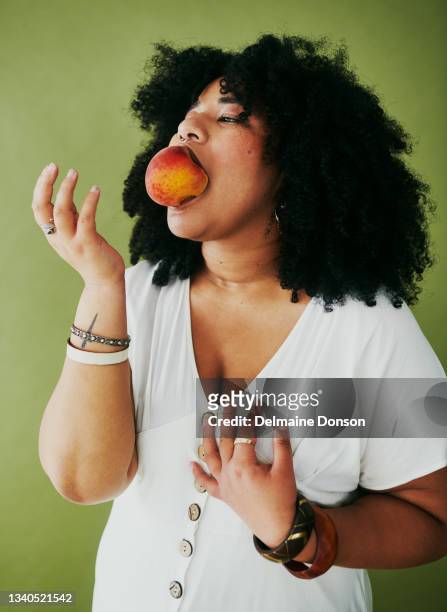 studio shot of a young woman eating a nectarine against a green background - plus size model fitness stock pictures, royalty-free photos & images