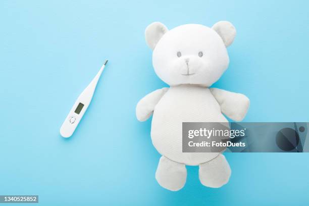 White teddy bear and digital thermometer on light blue table background. Pastel color. Closeup. Temperature measuring of children. Healthcare concept. Top down view.