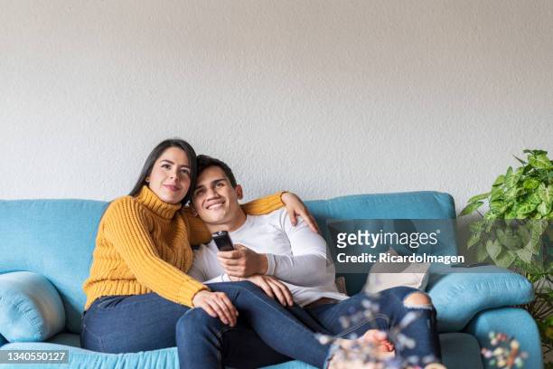 latin couple are sitting on the sofa at home watching television - young couple watching tv stock pictures, royalty-free photos & images
