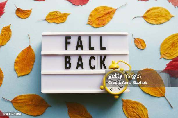 yellow retro alarm clock, autumn red and orange leaves over blue background. fall back, daylight saving time concept. - daylight saving time foto e immagini stock