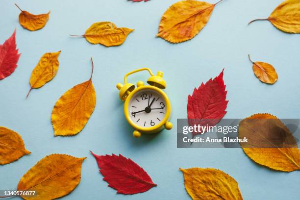 yellow retro alarm clock, autumn red and orange leaves over blue background. fall back, daylight saving time concept. - fall back fotografías e imágenes de stock