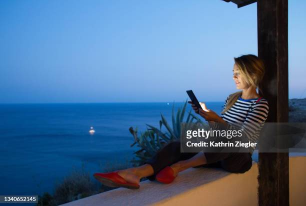 woman reading on digital tablet at dusk, sitting on terrace with sea view - digital storytelling stock pictures, royalty-free photos & images