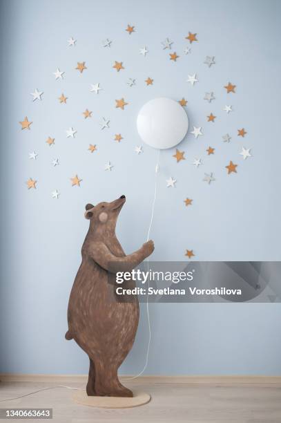 a wooden decorative bear stands on two legs and holds onto a wire from a wall lamp resembling a full moon. a fabulous interior composition of a bear and a starry sky on the wall of a children's room. - blue bear stock-fotos und bilder