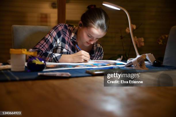 teenager doing paint by numbers art at a dining table in low light in the evening, under a portable desk lamp and wearing wireless in-ear headphones. - angle poise lamp foto e immagini stock