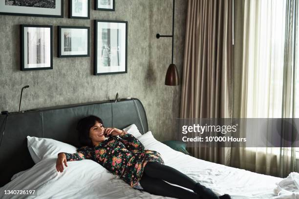 Cheerful young woman relaxing on hotel bed using phone